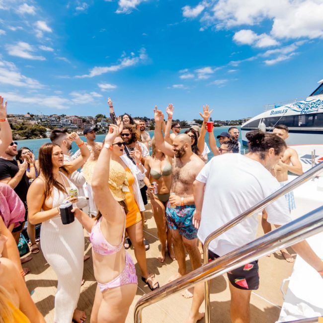 A group of people in swimwear enjoys a sunny day on a private yacht charter in Sydney Harbour, some with hands raised, set against a background of blue skies, water, and nearby buildings.