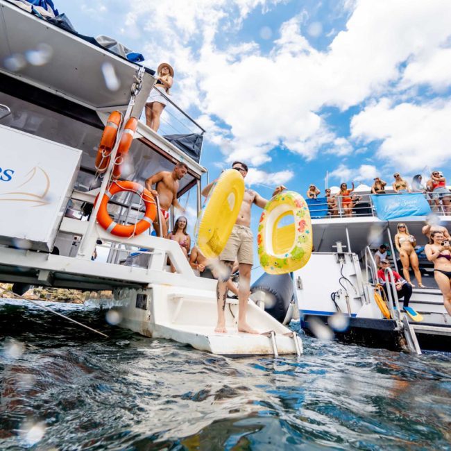 People are boarding a boat named "Whale One" with an inflatable yellow ring. Another boat with more people is docked nearby under a partly cloudy sky, setting the perfect scene for a private yacht charter on Sydney Harbour.