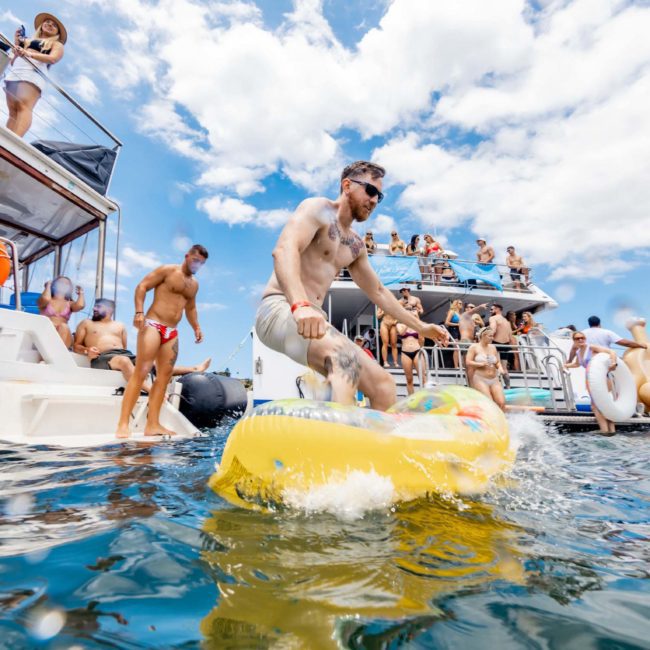 People enjoying a party on anchored boats, with a man riding an inflatable float in the water. Others are socializing and relaxing under a partly cloudy sky during a Sydney boat party hire.