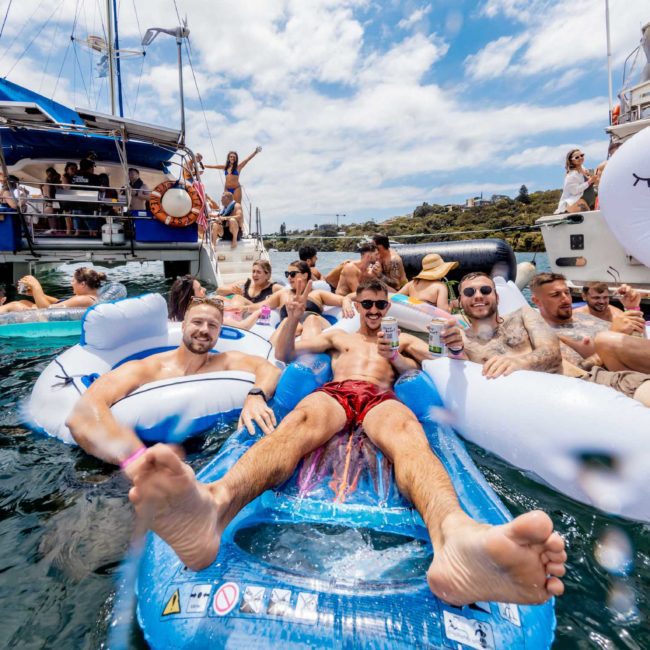 A group of people relax and socialize on inflatable floats in the water, surrounded by boats on a sunny day, with a luxury yacht hire Sydney providing an elegant backdrop.