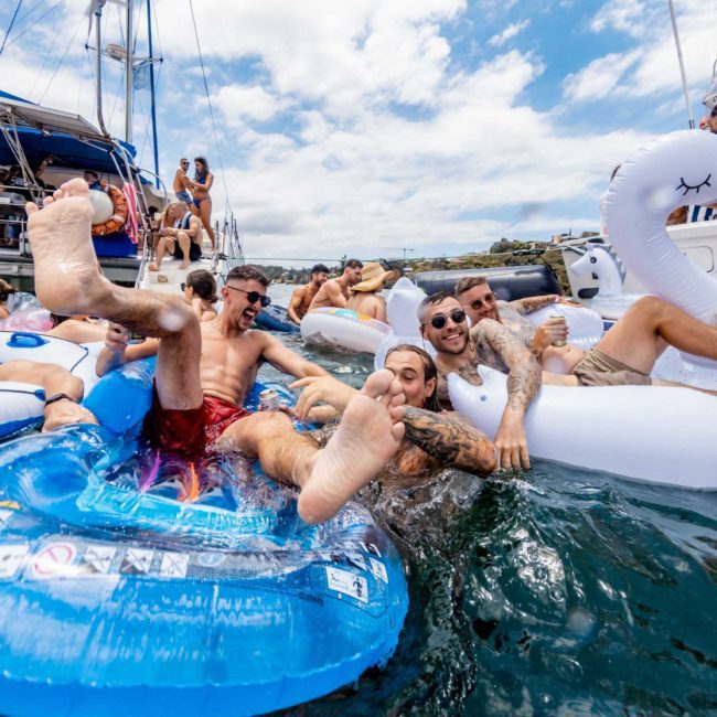 A group of people relax and have fun on inflatable floats near a catamaran party Sydney. Some smile at the camera, while others splash and interact with each other under a sunny sky.