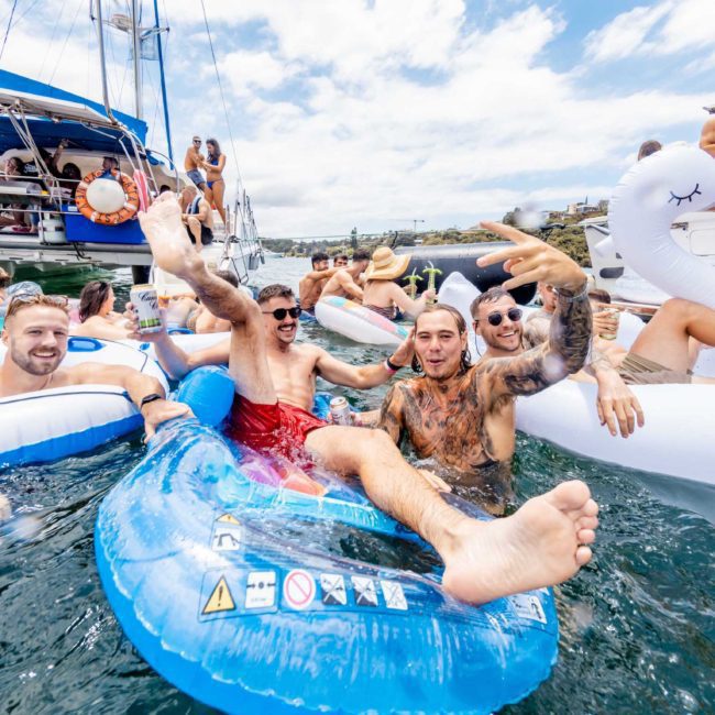 Group of people enjoying a party on water floats along a docked boat on a sunny day, surrounded by various inflatable toys. The scene epitomizes the elegance of a private yacht charter in Sydney Harbour.