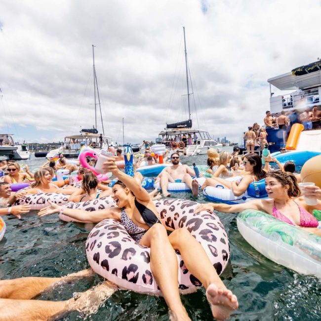 A large group of people enjoying a Sydney boat party hire on the water, floating on inflatables and swimming, with several boats in the background under a partly cloudy sky.