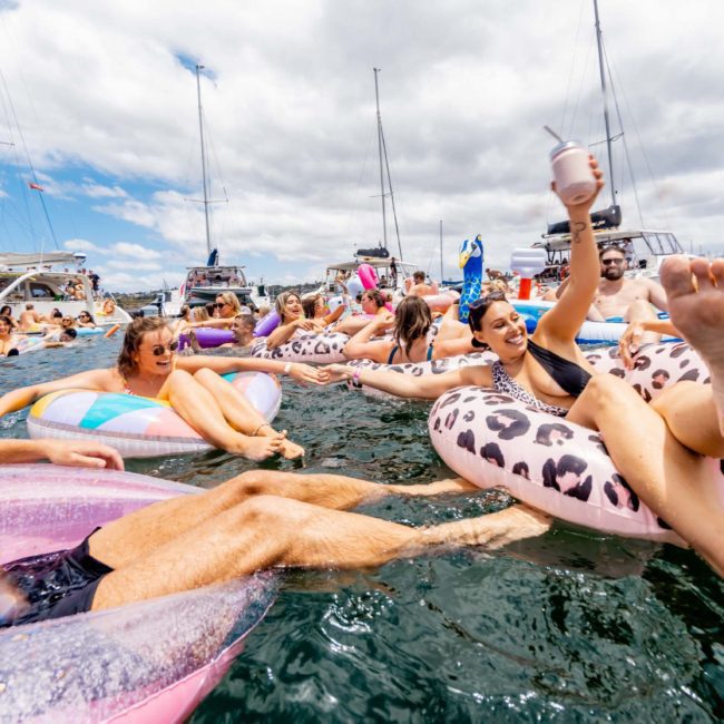 People relax and enjoy drinks while floating on inflatable tubes in the water, surrounded by boats on a sunny day. Ideal for those looking for a Sydney boat party hire or corporate boat events Sydney.