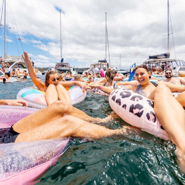 A group of people enjoy a sunny day on the water, floating on inflatable tubes and surrounded by boats. Some wave and smile, creating a festive atmosphere reminiscent of a Sydney boat party hire event.