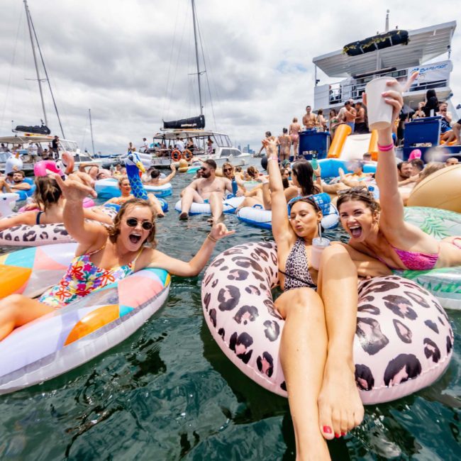 People relax on floaties in the water, raising drinks and smiling, with boats and more people in the background. The scene appears festive with a mix of colorful inflatable rafts, ideal for those looking into luxury yacht hire Sydney or DJ boat hire Sydney for their next event.
