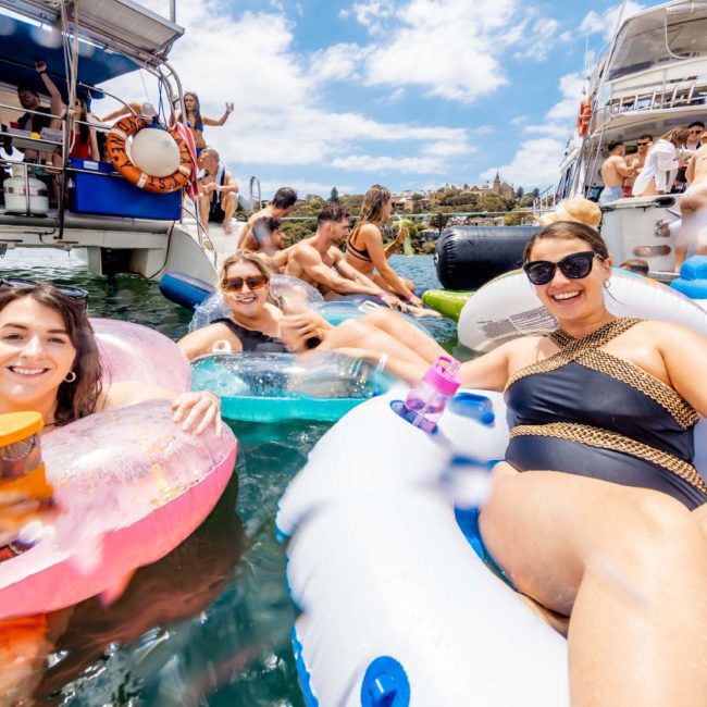 A group of people enjoying a sunny day on inflatable rafts in the water, with boats and more people in the background. One person holds a drink while others relax and smile, making it a perfect scene for a private yacht charter Sydney Harbour or DJ boat hire Sydney.