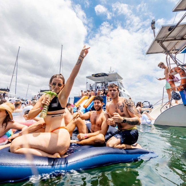 On a sunny day, a group of people relishes the water, some on a floating mat and others on a private yacht charter in Sydney Harbour. One person raises an arm while holding a beverage, adding to the festive atmosphere. The background showcases multiple boats cruising by.