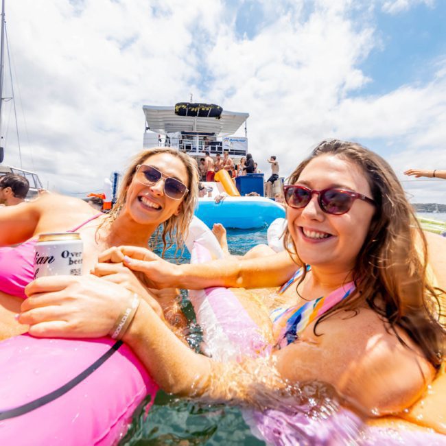 Two women in swimsuits and sunglasses hold drinks while relaxing on inflatable floaties in the water. A luxury yacht hire and people are in the background on a sunny day.