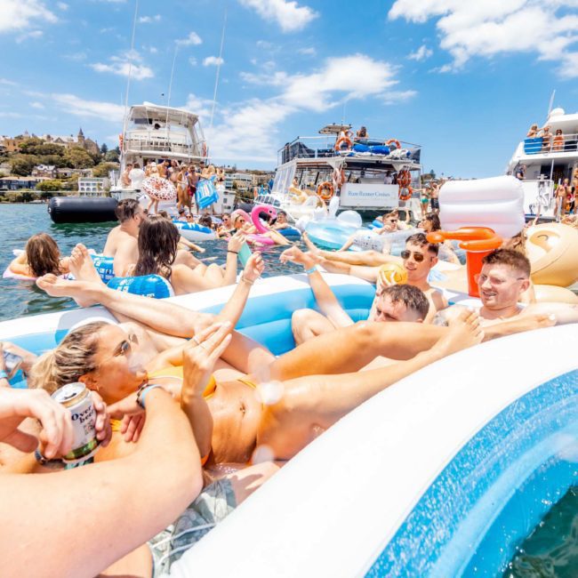 A group of people enjoying a sunny day in inflatable pools and floats on the water, surrounded by boats and other people, partaking in the fun of a Sydney boat party hire.