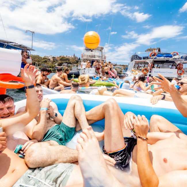 A group of people relax in an inflatable pool on water surrounded by boats and floats, under a partly cloudy sky, enjoying a Sydney boat party hire.