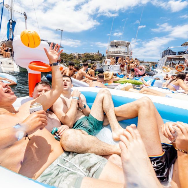 A group of people in swimsuits relax and have fun on inflatable rafts and boats in a crowded water area under a clear sky, enjoying the vibe reminiscent of a luxury yacht hire Sydney.