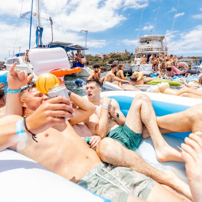 People relaxing and socializing on inflatable tubes in the water, with boats and more people in the background on a sunny day. Some are holding drinks, and one person is being playfully sprayed with water. It looks like a perfect setting for a Catamaran party in Sydney.