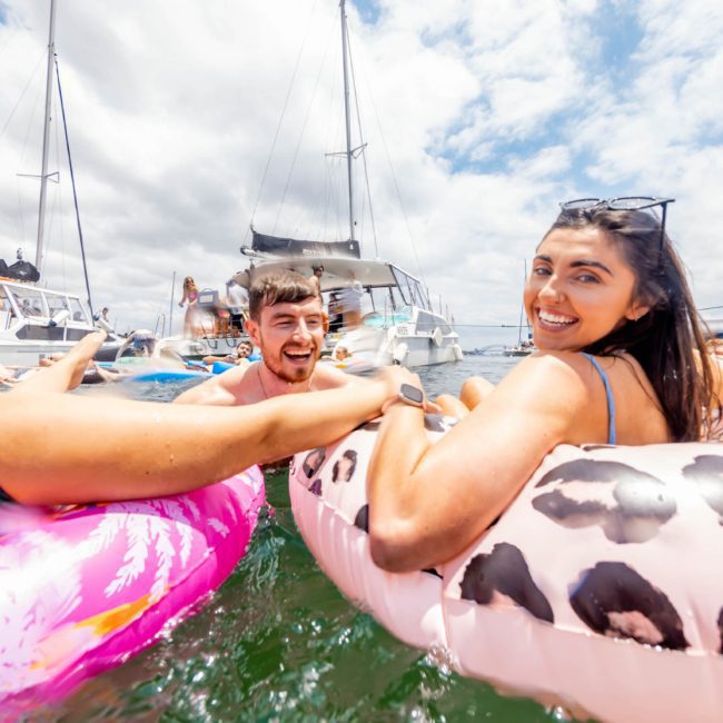 Three people are smiling and relaxing on inflatable floaties in the water near a marina, with several boats, including a luxurious catamaran party Sydney, in the background under a partly cloudy sky.