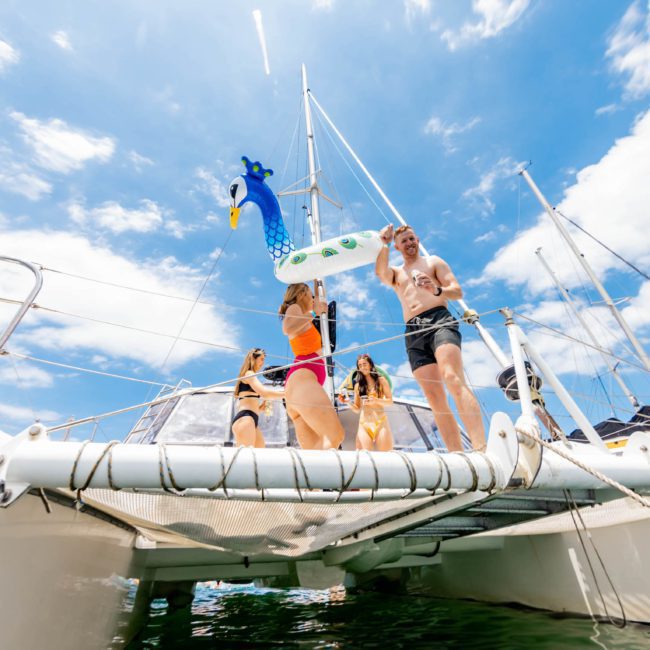 People on a catamaran enjoy a sunny day holding inflatable toys, with a few individuals standing near the boat's netted area and a clear blue sky in the background, making it the perfect Catamaran party Sydney experience.