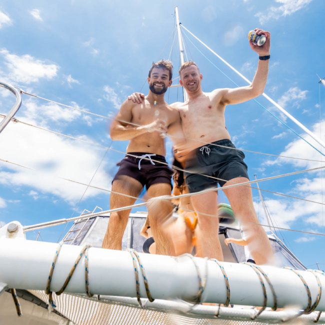 Two men in swim trunks stand on the deck of a boat, smiling and holding drinks, with a clear blue sky and other boats in the background—a perfect scene for a Sydney boat party hire.