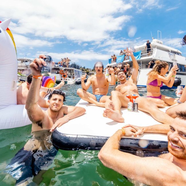 Group of people enjoying time on a floating platform and inflatable unicorn in the water, with boats and more people in the background on a sunny day, perfect for a Sydney boat party hire.