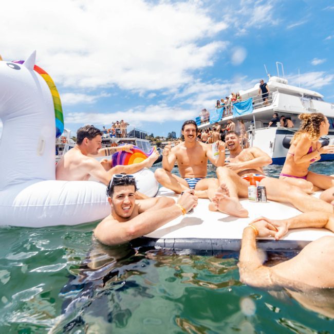 People relaxing on inflatable floats, including a unicorn float, in the water on a sunny day with boats and others in the background. A luxury yacht hire Sydney can be seen adding elegance to the scene.