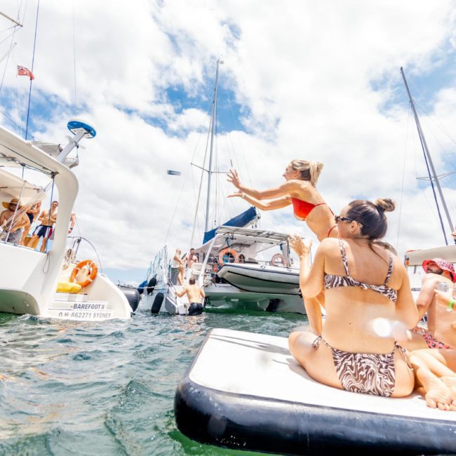 People are enjoying a sunny day on boats and inflatable floats in the water. One person in a red swimsuit is mid-jump into the water, while others watch and relax, making it the perfect scene for a Catamaran party Sydney.