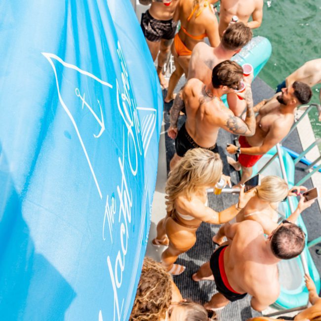 Group of people in swimwear socializing on a docked boat with a blue "The Yacht Club" banner visible to the side above a body of water, enjoying a Sydney boat party hire.