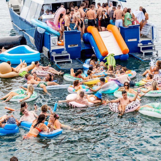 A large group of people enjoy floating on inflatables and swimming near a docked boat in a body of water, with some using an inflatable slide from the boat, reveling in the ultimate luxury yacht hire Sydney experience.