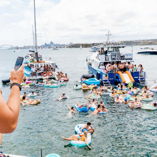 A man using a smartphone overlooks a group of people in floats and inflatable toys gathered around boats in a busy waterfront scene, possibly enjoying a Sydney boat party hire.