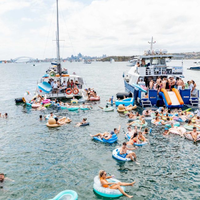 People enjoy a sunny day on the water, floating on various inflatables near two boats connected by slides, with the city skyline visible in the background. Experience the ultimate fun with Sydney boat party hire or treat yourself to a luxury yacht hire Sydney for an unforgettable day.