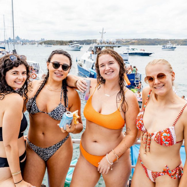 Four women in swimsuits are posing for a photo on a luxury yacht with a scenic view of the water, boats, and skyline in the background, epitomizing the perfect Sydney boat party.