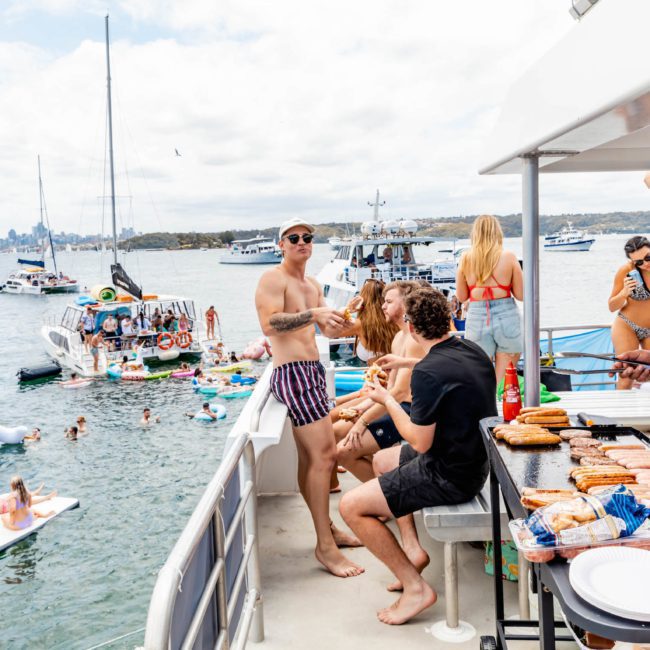 A group of people enjoying a lively catamaran party in Sydney Harbour with others in the water and on nearby boats. Food is being grilled on the deck while attendees socialize and relax on inflatable floats, creating a perfect day of fun and relaxation.