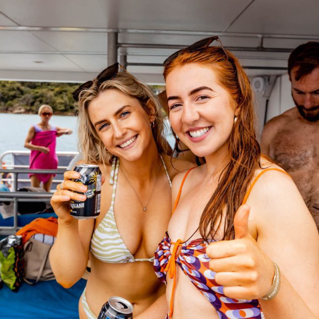 Two smiling women in swimsuits hold drinks on a boat, enjoying a lively catamaran party in Sydney. One gives a thumbs-up. Other people are visible in the background, including a man adjusting his swim trunks.