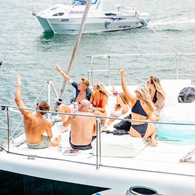 A group of people in swimwear are sitting and raising their arms on a yacht in a body of water, enjoying a Sydney boat party hire. Another boat is seen in the background.