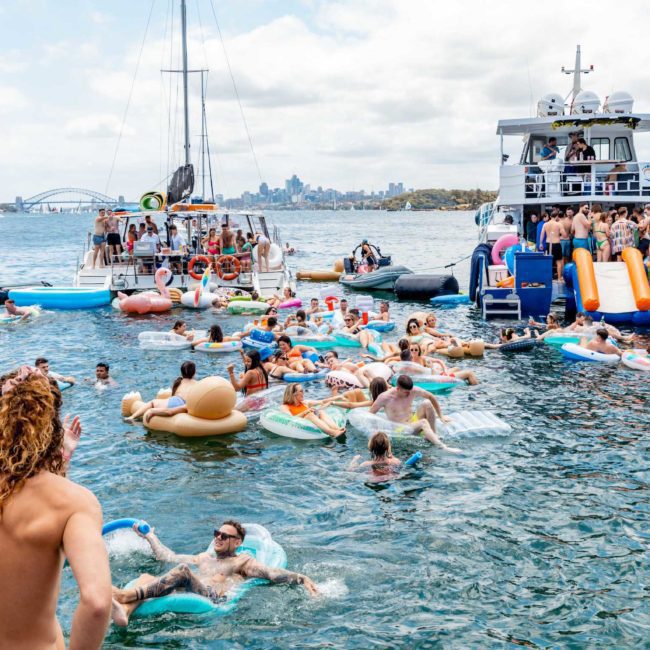 A large group enjoys a catamaran party Sydney, swimming and floating on inflatables in the water, with multiple boats and a city skyline in the background.