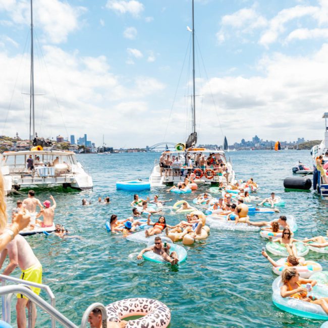 A crowd of people on inflatable floats and boats enjoy a sunny day on the water. Some are swimming, while others relax on luxurious yachts docked nearby, perfect for corporate boat events Sydney. The skyline of a city can be seen in the background.