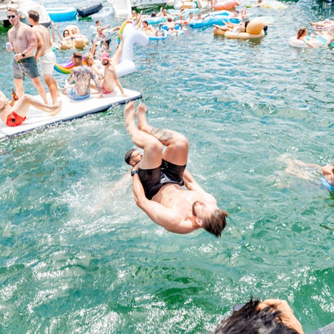 Two individuals are in mid-air after jumping into a body of water, surrounded by people on boats and inflatables, enjoying a sunny day at an epic Sydney boat party hire.