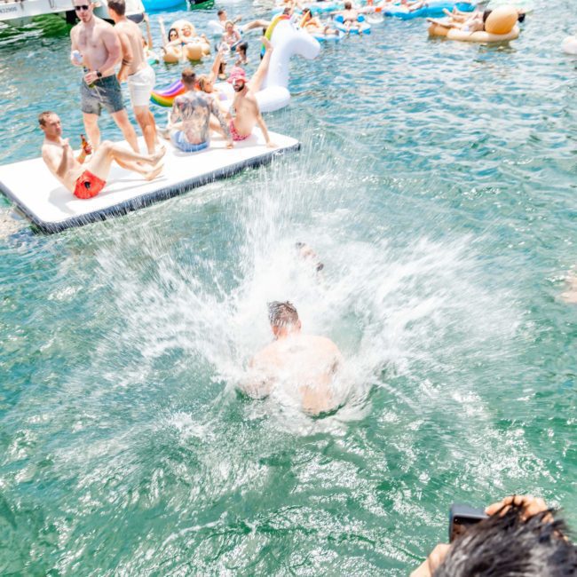 People enjoying a sunny day on boats and inflatable rafts in the water. One person is mid-dive, creating a large splash. The scene is lively with a mix of activities, accentuated by the elegance of luxury yacht hire Sydney.