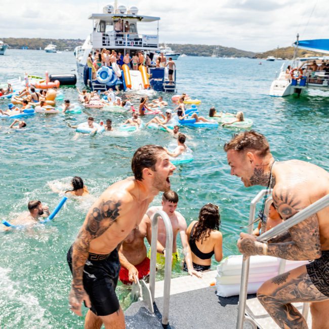 People are enjoying a sunny day on the water with floats and boats. Two tattooed men are smiling and interacting at the edge of a boat ladder, typical of a lively Sydney boat party hire. Other boats and swimmers are visible in the background, perfect for corporate boat events in Sydney.