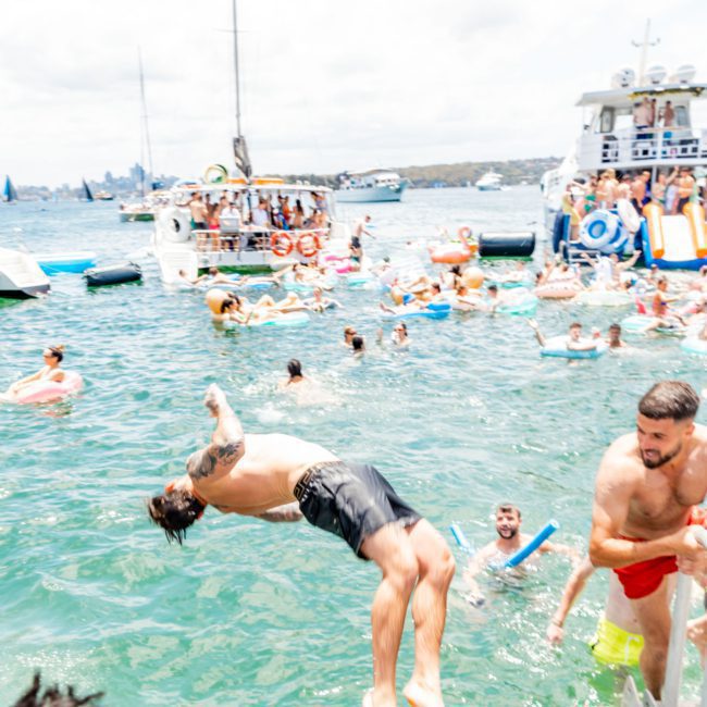 A person performs a backflip into the water as others swim and relax, with boats and inflatables in the background, epitomizing a lively Sydney boat party hire.