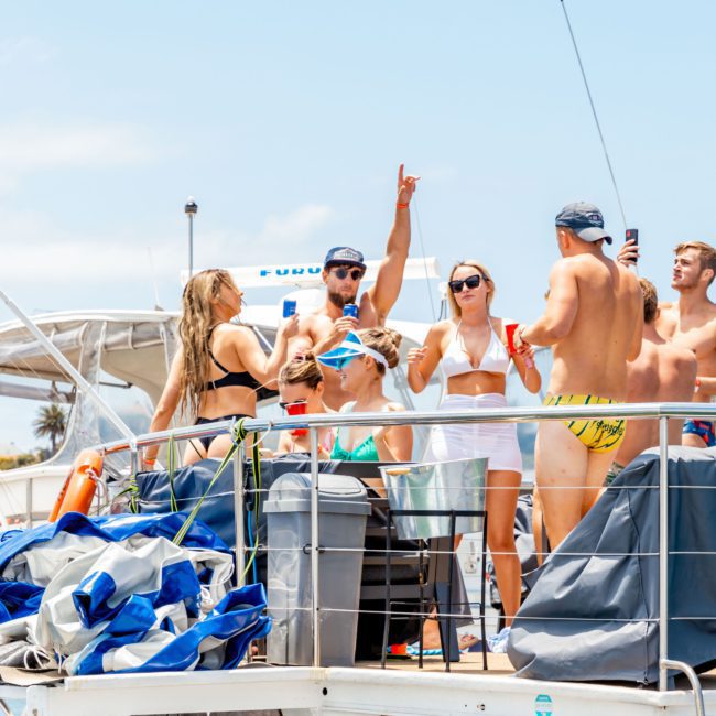 A group of people on a boat are partying, with some holding drinks and others dancing. The boat, a catamaran party Sydney vessel, has lifebuoys labeled "Rum Runner" and various equipment on board.