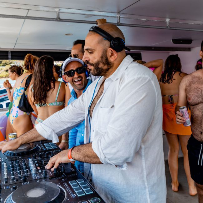 A man wearing headphones DJs from a console on a boat surrounded by people in swimwear, making it an unforgettable Sydney boat party hire.