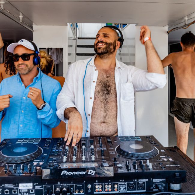 A man DJs on a Pioneer DJ controller with headphones on, while another man in sunglasses stands beside him, also wearing headphones. In the background, a shirtless person enjoys the vibrant atmosphere of a Sydney boat party hire.
