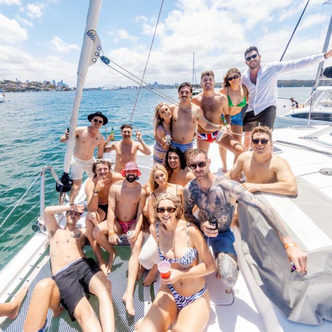 A group of people in swimsuits smiles and poses on a sailboat under clear skies with water and other boats visible in the background, making the most of their Sydney boat party hire.