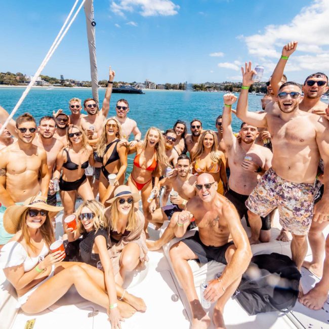 A group of people in swimwear are gathered on a Private yacht charter Sydney Harbour, enjoying a sunny day, holding drinks, and posing for a photo near a coastal area.