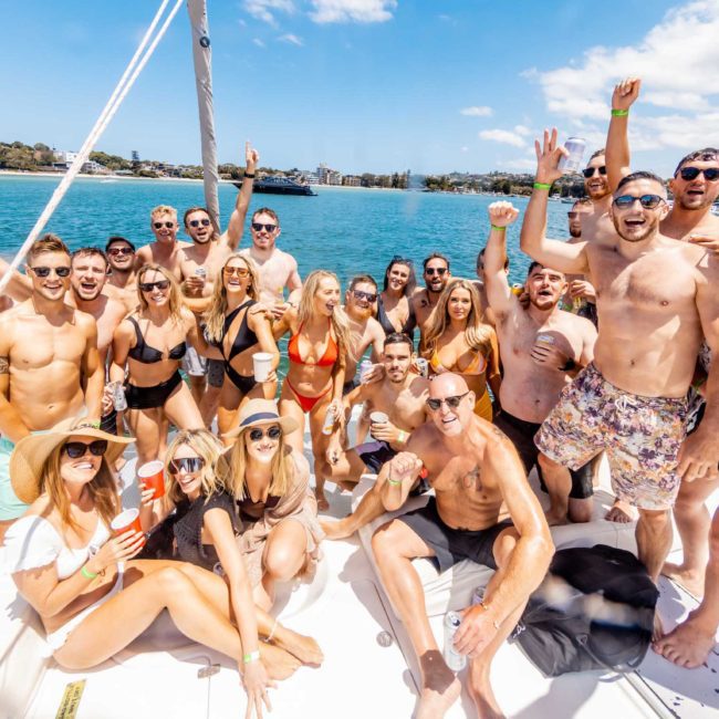 A group of people in swimwear enjoying a sunny day on a sailboat, celebrating with drinks. With the stunning Sydney coastline as their backdrop, this vibrant Catamaran party Sydney experience showcases pure joy and excitement.