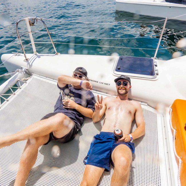 Two people relax on a netted area of a catamaran, one wearing a hat and shaded glasses, the other shirtless. Both appear to be enjoying drinks with a view of Sydney Harbour in the background, making it an ideal scene for a private yacht charter or corporate boat event.