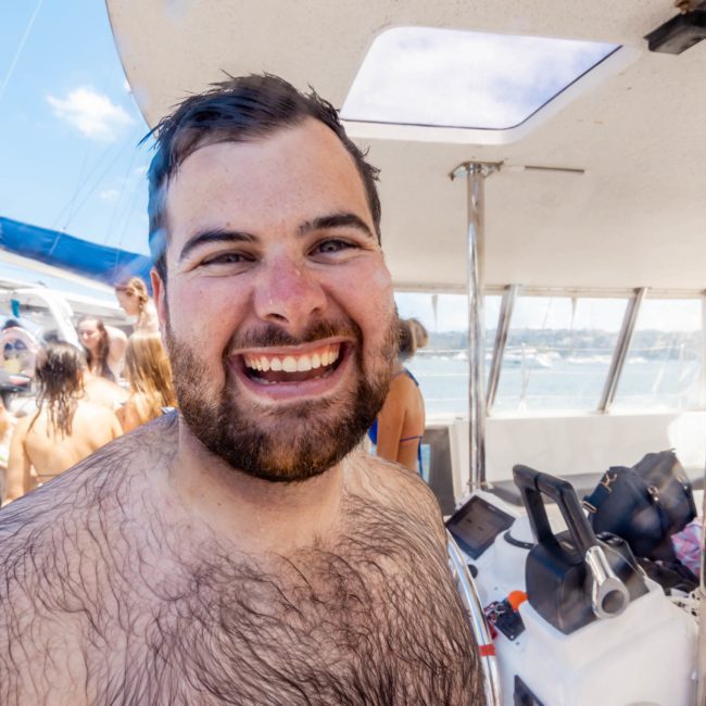 A man with a wet beard and chest smiles on a boat with people in swimsuits in the background, enjoying a lively Sydney boat party hire.