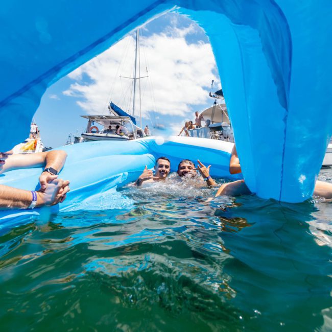 People in the water hold onto a large blue inflatable near boats on a sunny day, enjoying their luxury yacht hire Sydney experience.