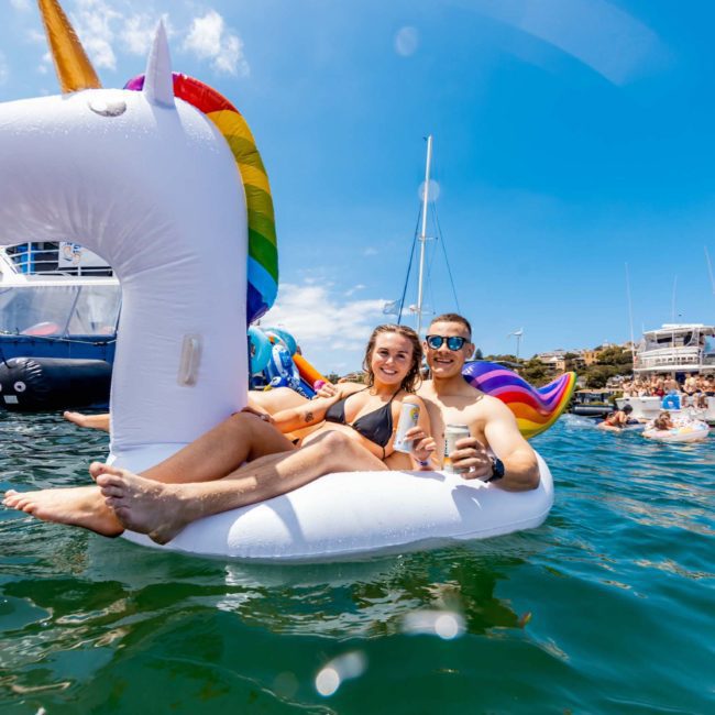 Two people on an inflatable unicorn float in the water, holding drinks, with boats and several other people enjoying a Sydney boat party hire on a sunny day.