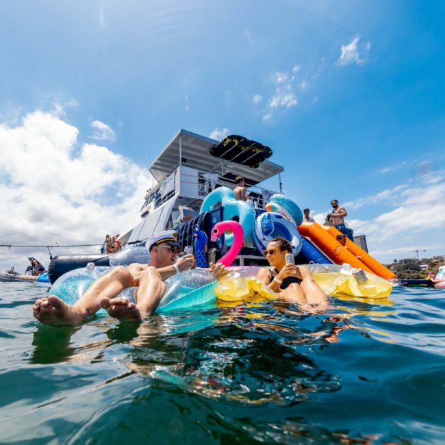 People relax on inflatable floats in the water near a boat with a slide during a lively Catamaran party Sydney. The sky is clear with a few clouds, and other boats are visible in the background.