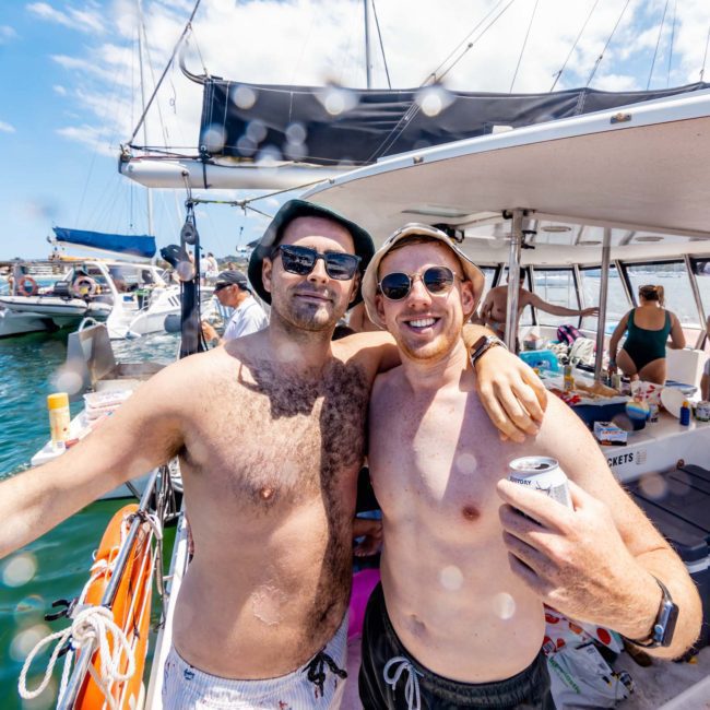 Two men with arms around each other, smiling on a sailboat with other people in the background. One holds a can and wears sunglasses and a hat. Other boats and water activities are visible, showcasing the perfect day for a luxury yacht hire in Sydney.