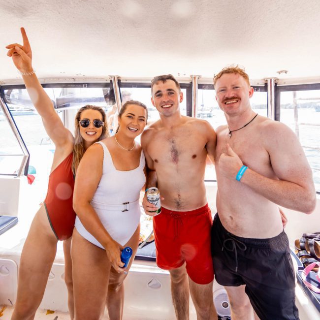 Four people in swimsuits smiling and posing for a photo on a catamaran party in Sydney. One person on the left is pointing upwards while another adjusts their swimsuit.